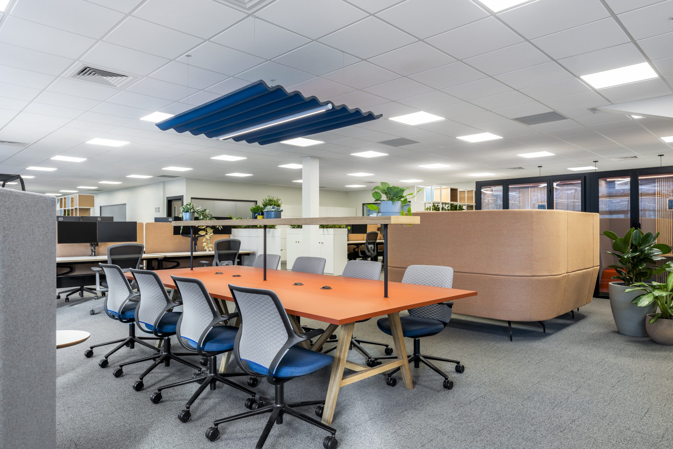  Open-plan office interior featuring a long orange collaboration table surrounded by blue and grey ergonomic chairs